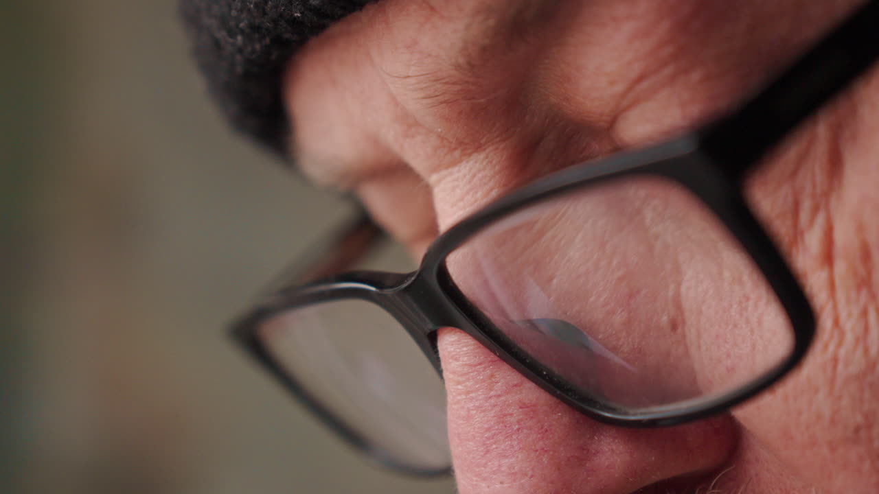 Scatto macro estremo del viso e degli occhiali di un uomo mentre si concentra a scrivere in un diario. La luce fioca e l'inquadratura ravvicinata enfatizzano l'intensa concentrazione, i pensieri segreti e la profonda emozione.