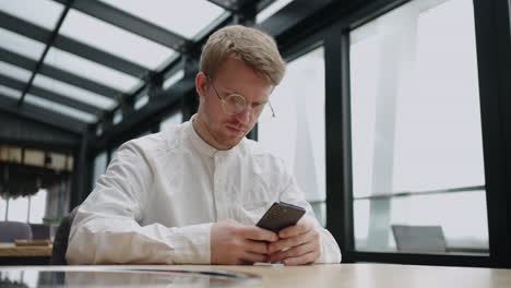 A-young-man-with-glasses-with-a-mobile-phone-in-his-hands-looks-at-the-screen-and-prints-a-message.-Hipster-in-round-glasses-writes-a-message-on-his-phone