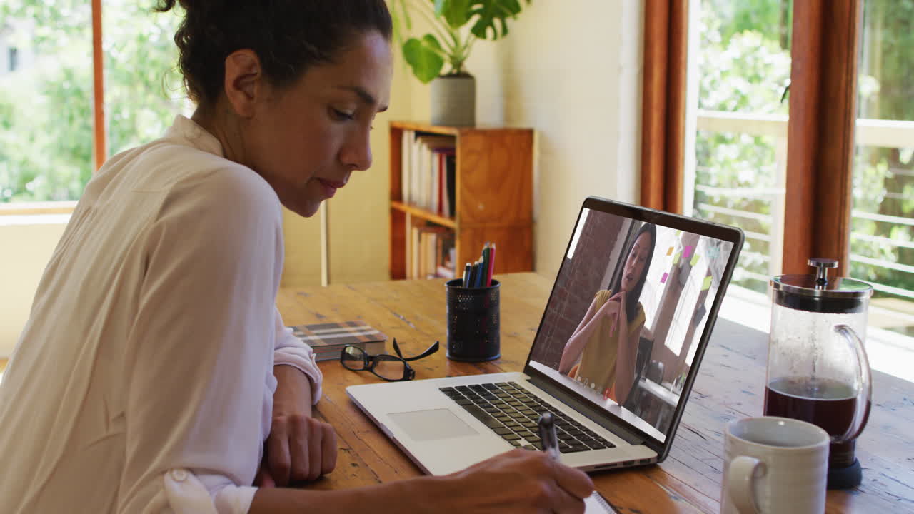 African american woman taking notes while having video call with female colleague on laptop at home