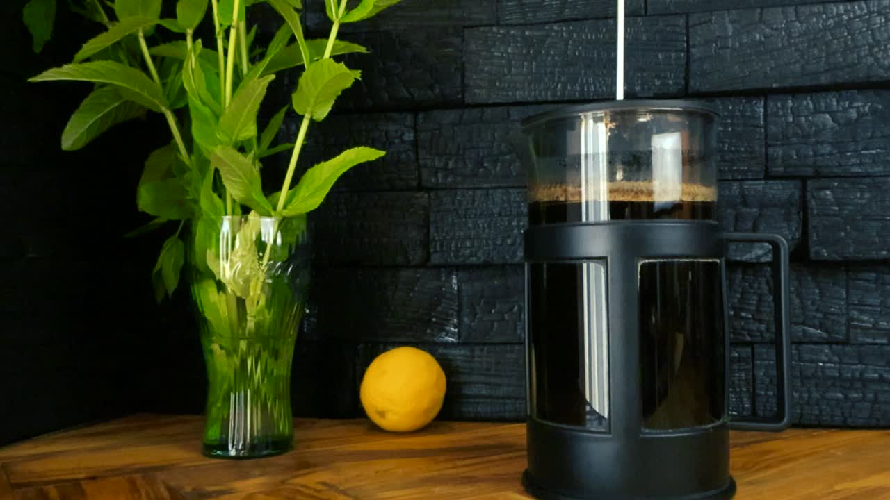 Gorgeous video footage of a french coffee press glass jar on an olive wood brown colored table with burned yakisugi design wall in the kitchen background. Yellow lemon and green peppermint plants seen