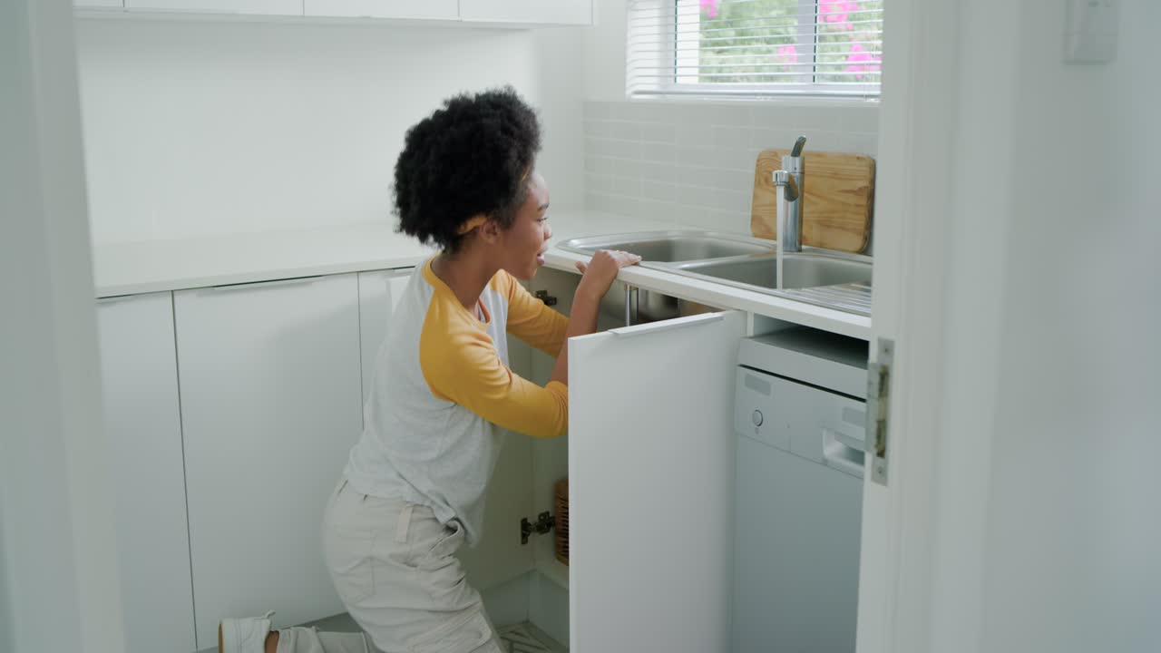 Woman kneeling opening cabinet under sink in kitchen, inspecting plumbing and using toolkit
