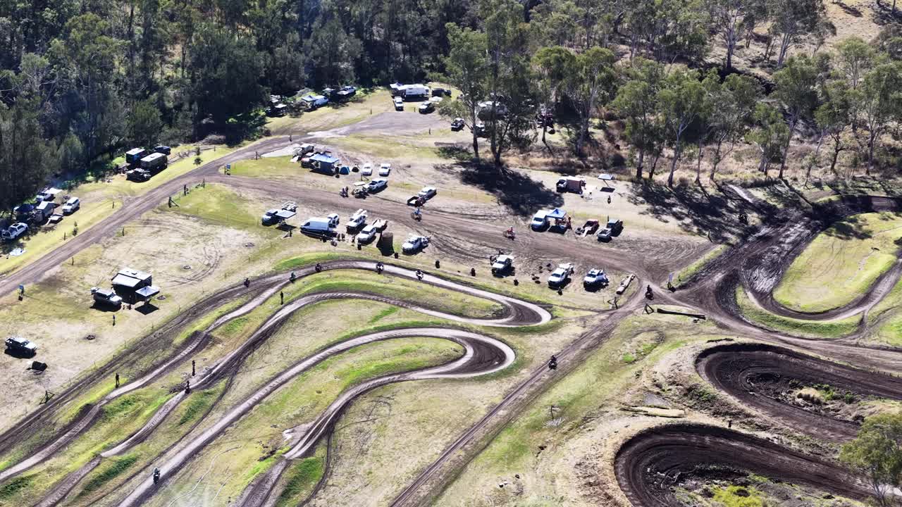 Drone captures winding motocross track, vehicles, and forested surroundings in bright daylight, steady overhead shot