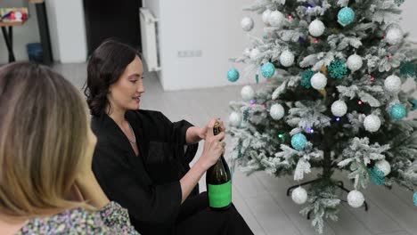 Two women celebrate holidays by opening a bottle of champagne next to a Christmas tree