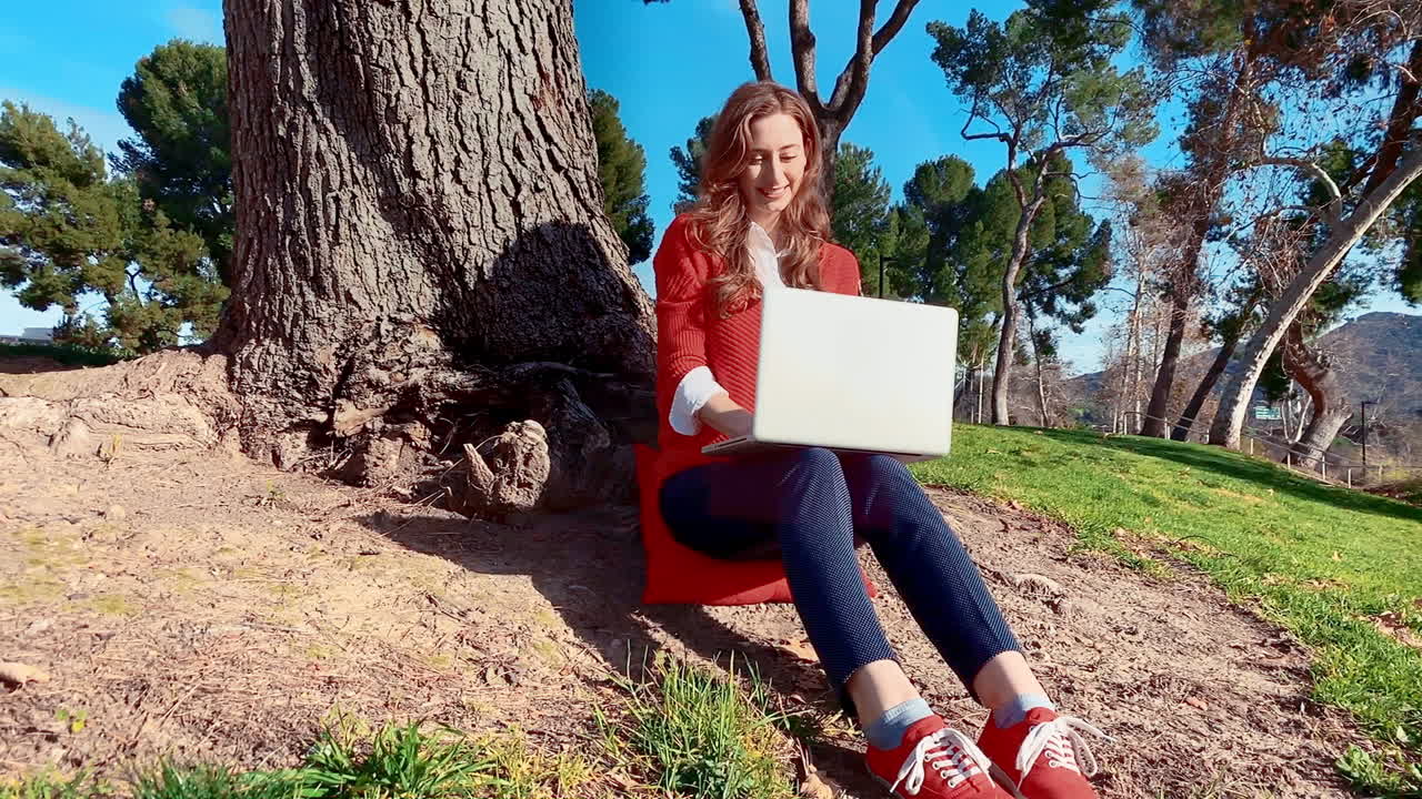 una mujer feliz, atractiva, joven y caucásica en el parque, escribiendo en su computadora portátil debajo de un árbol