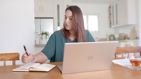 mujer biracial ocupada de tamaño plus escribiendo notas y usando portátil, trabajando desde casa, en cámara lenta