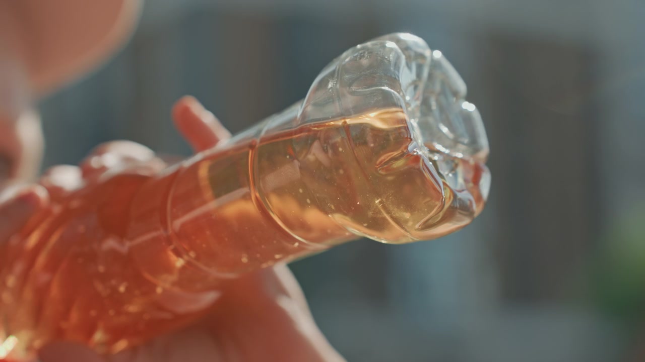 Una mano tiene una bottiglia di plastica trasparente riempita con liquido dorato, inclinandola per mostrarne il contenuto. L'attenzione rimane sull'interazione tra il contenitore trasparente e il suo fluido dalle sfumature arancioni.