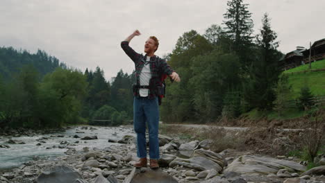 Male-hiker-standing-at-rocky-river-shore-in-mountains.-Redhead-man-raising-hands