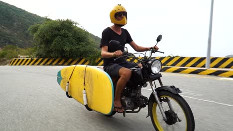 motorcyclist with yellow helmet and kitesurf board fasten to motorcycle drives with flip-flops