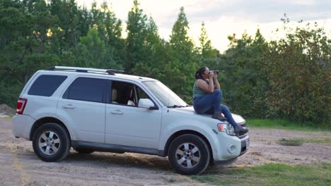 mujer con cámara dslr en sus manos, sentada en el capó de su coche todoterreno 4x4, tomando fotografías al aire libre durante la puesta de sol