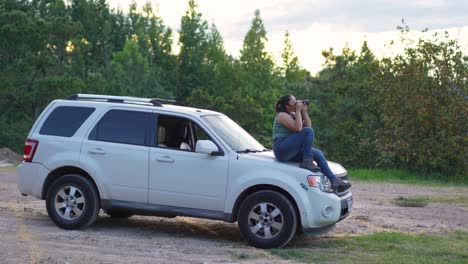 woman with dslr camera in her hands, sitting on the hood of her 4x4 suv offroad car, taking photographs outdoors during sunset