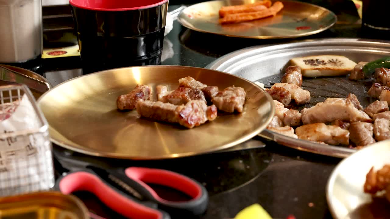 Tongs transfer grilled pork belly onto a plate at a lively Korean barbecue restaurant table