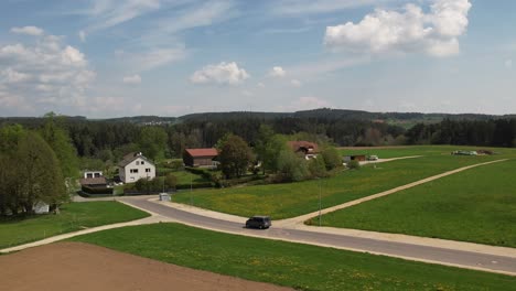a car drives on a rural road in upper palatinate, passing green fields and scattered homes, with wind turbines visible on the distant hills.