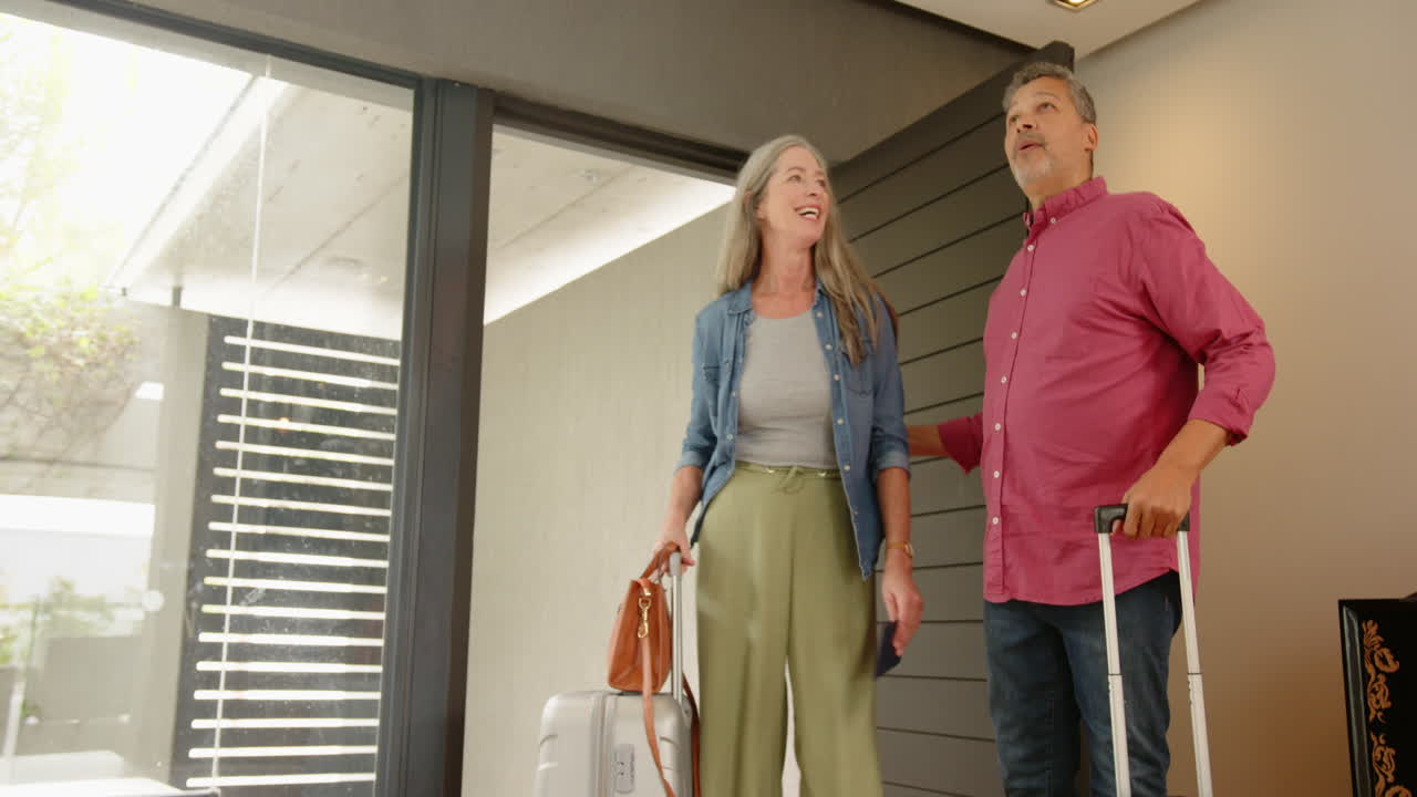 Arriving home, multiracial senior couple with luggage smiling and pointing, copy space