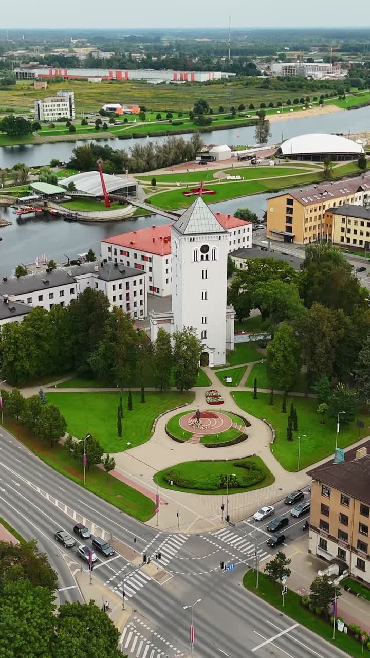 Vertical aerial view of Jelgava, Latvia—Mitava Bridge spans Lielupe River, framed by Holy Trinity Tower, civic buildings, and green parkland in city center