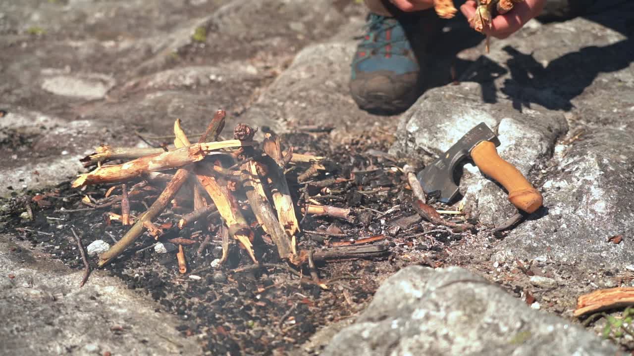 Person adding dry natural branches to freshly lit bonfire in rocky firepit - Smoke and flames coming from firewood - Sunny closeup nature clip in slow motion