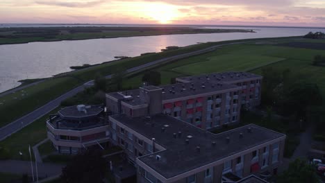 Reveal-shot-of-Avondrust-Makkum-next-to-dyke-and-ijsselmeer-with-sunset,-aerial