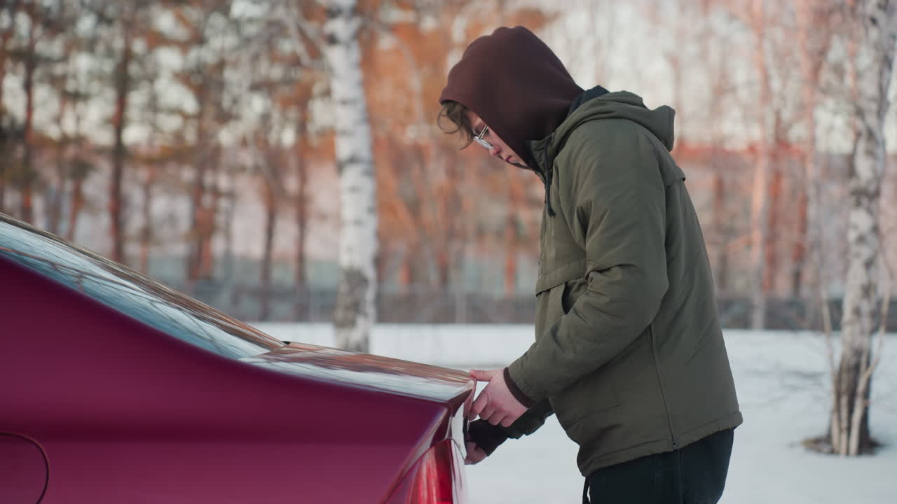 Side view of teenager wearing winter jacket and hoodie closing red car boot, cold outdoor setting with snow on ground, blurred trees in background, hands on handle with reflection visible on surface