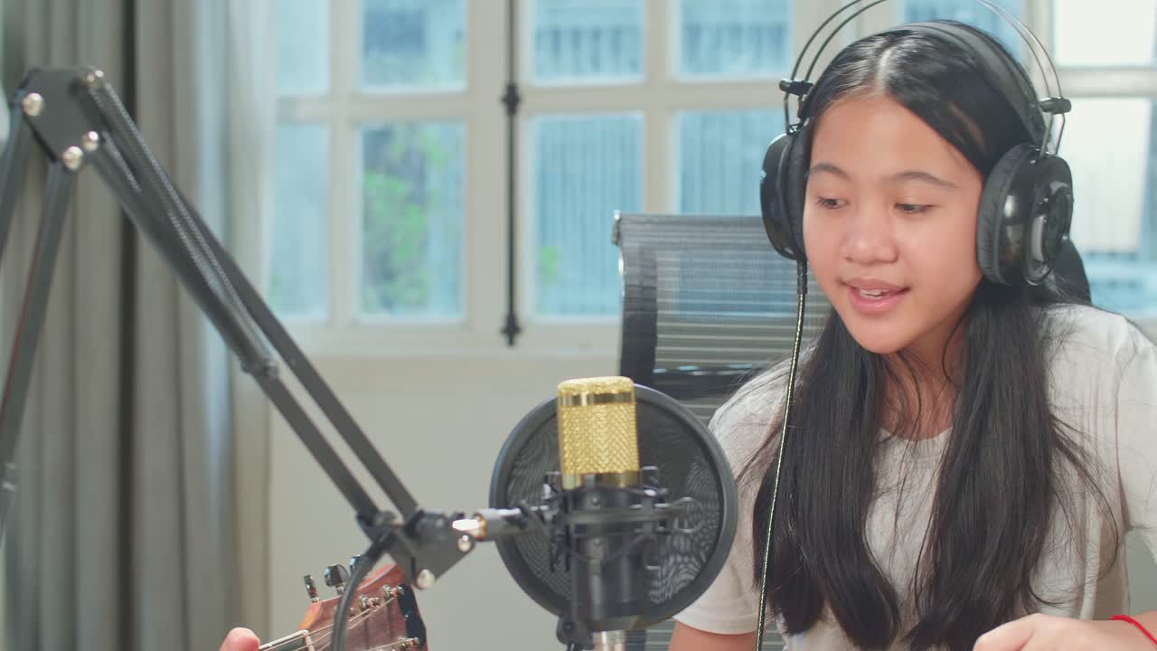 Asian Kid Girl In Headphones Singing Into Microphone While Boy Playing The Guitar During Online Livestream In Podcast Studio