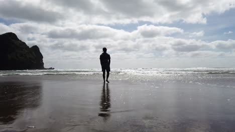 man walks towards the sea, individual strolling on beach, hands in pockets, slow