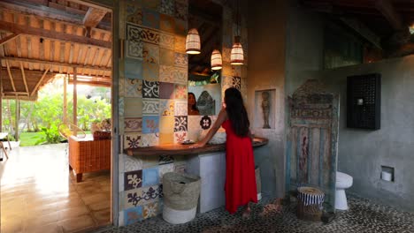 Mujer-Asiática-Con-Un-Maxi-Vestido-Rojo-Mirando-El-Reflejo-Del-Espejo-En-El-Baño-Temático-Joglo