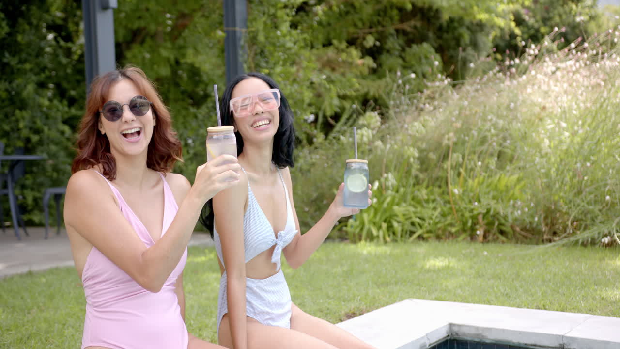 Two biracial female friends holding drinks by pool at home, copy space