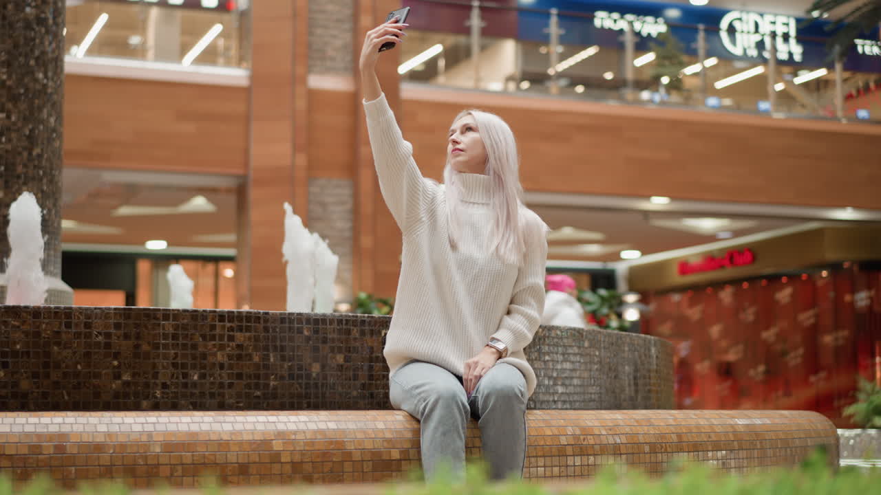 Fashionable shopper sits on tiled bench beside indoor mall fountain, raises smartphone high to capture water jets and ambient lights, people strolling past retail storefronts creating media content
