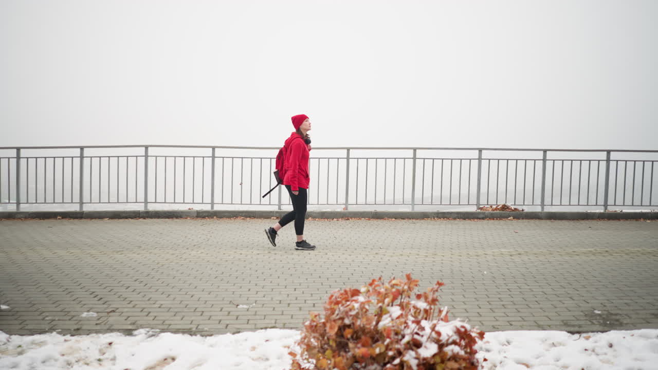 Side view of athlete in red sweater with bag over shoulder walking near iron railing on snow-dusted ground with scattered leaves, surrounded by a foggy atmosphere creating a calm and serene setting