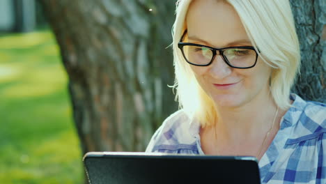 attractive young woman in glasses uses a tablet sits in a park near a tree beautiful light before su