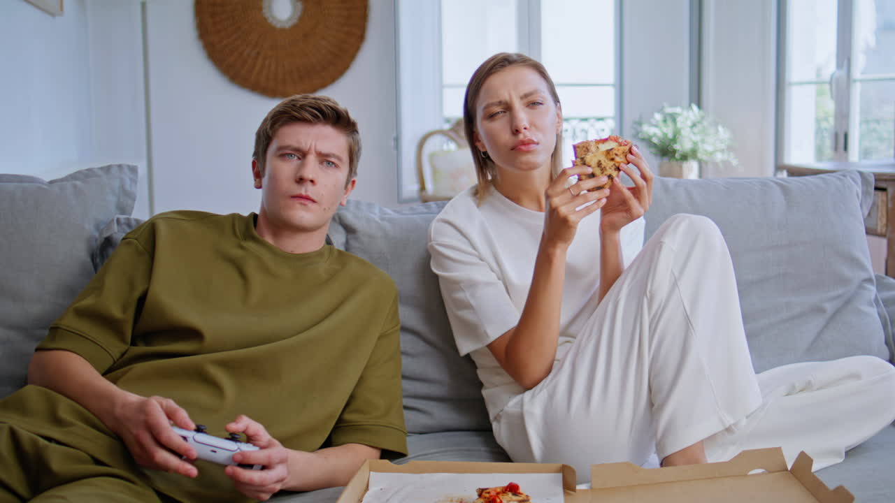 Homie lovers playing console sitting sofa together. Young woman eating pizza
