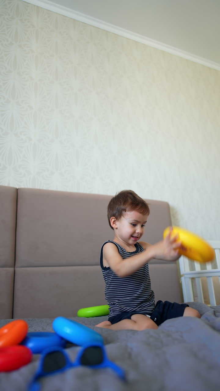 Lovely baby boy put a ring from toy pyramid on his head. Little toddler plays on a big bed at home. Vertical video.