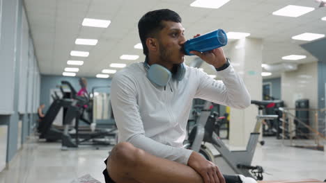 young man seated on bench in bright indoor gym drinking from blue bottle with wireless headphones around neck while others exercise on treadmills in blurred background under ceiling lights