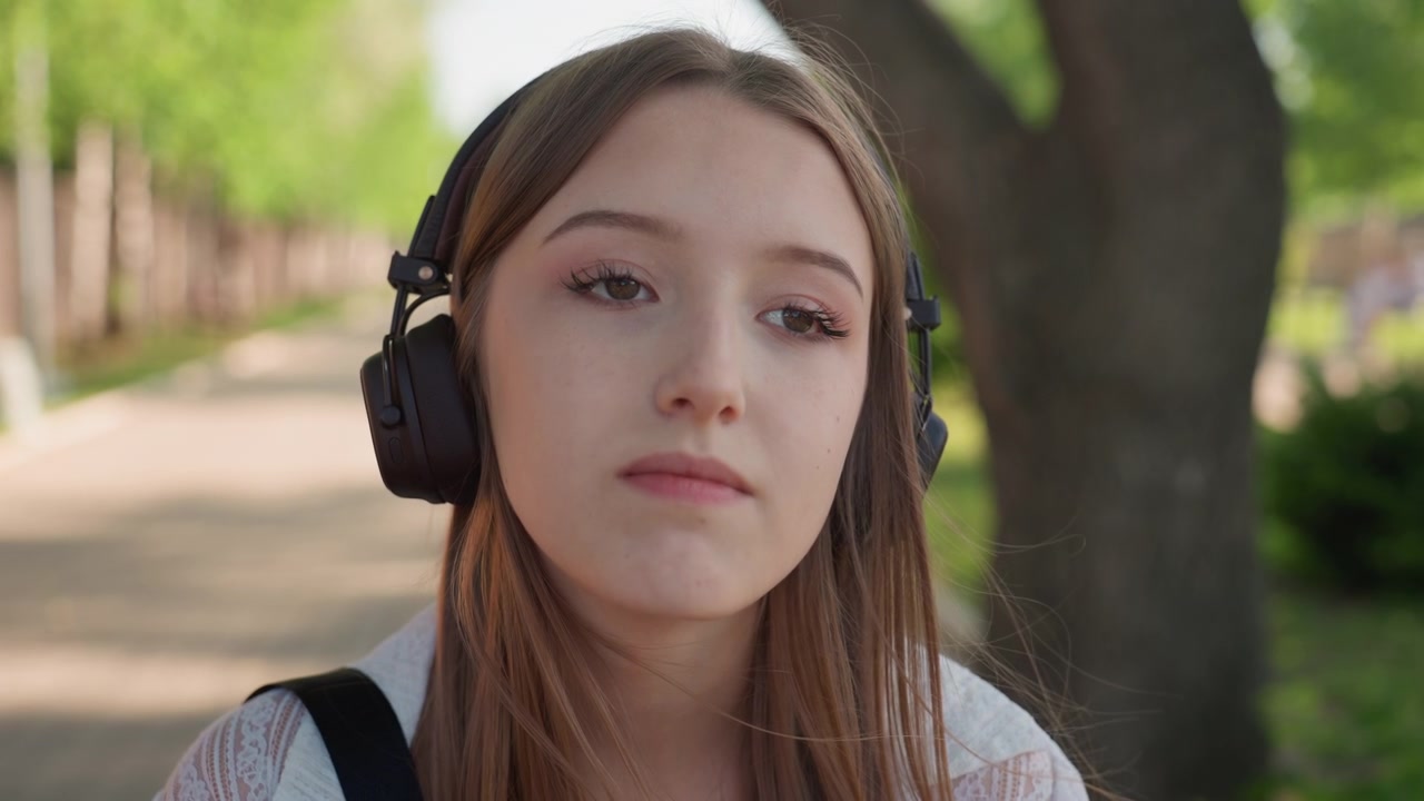 Primo piano di giovane donna che indossa cuffie e porta uno zaino, in piedi all'aperto su un sentiero con alberi sullo sfondo. La scena è illuminata dolcemente dalla luce naturale del giorno, creando toni caldi.