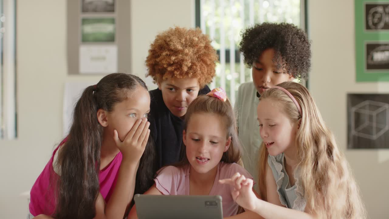 Verschiedene Kinder versammeln sich um ein Tablet auf einem Tisch im Klassenzimmer, das rechte Mädchen tippt und teilt den Fokus
