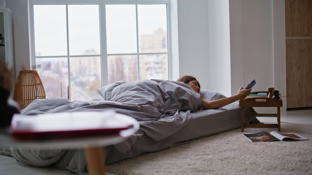 Morning lady checking time cellphone covering blanket continue napping in bed