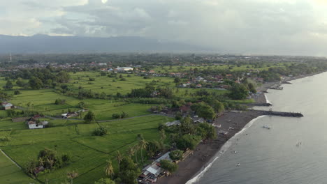 aerial view of a coastal village in bali, with people on a black sand beach, boats by the jetty, and green rice fields backed by distant mountains and captured at sunset