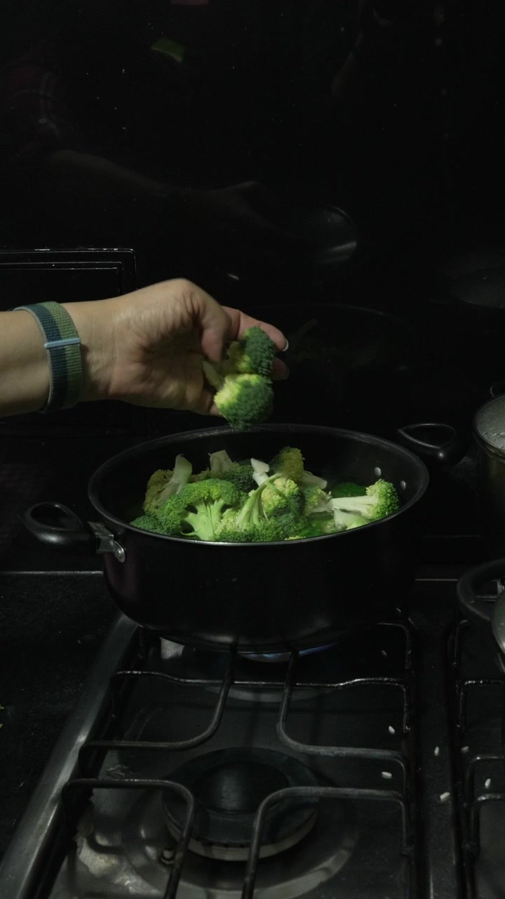 Una singola mano rilascia pezzi di broccoli freschi in una pentola di acqua bollente su un fornello a gas in una cucina domestica buia, utile per la cucina sana o contenuti di ricette