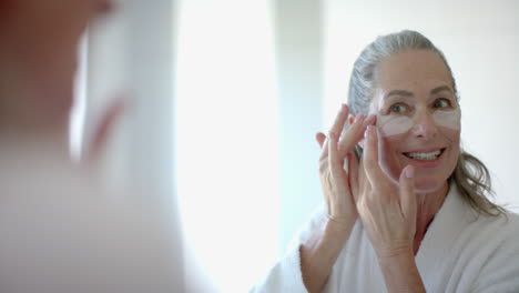 applying under-eye patches, mature woman smiling and looking at mirror in bathroom