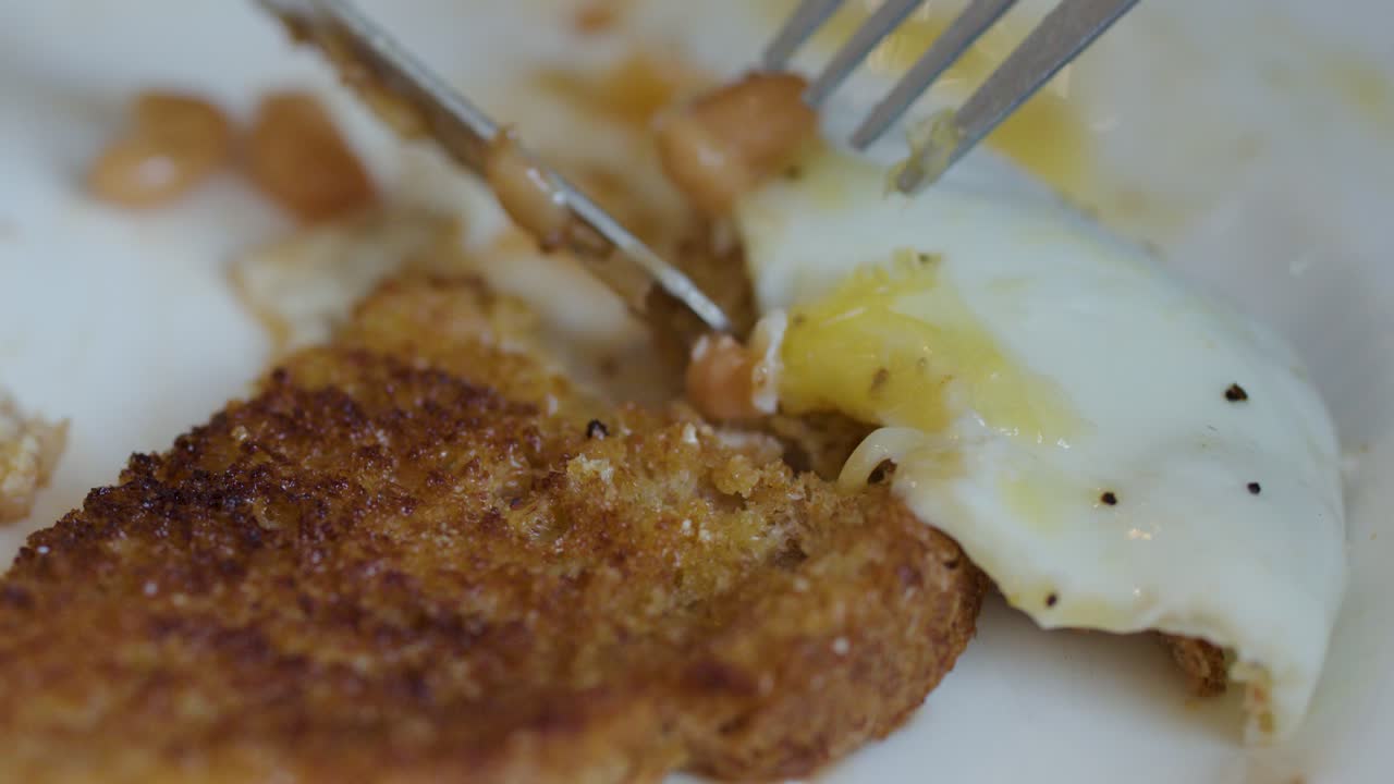 Close-up of toast, egg, and beans being cut with fork and knife under natural lighting
