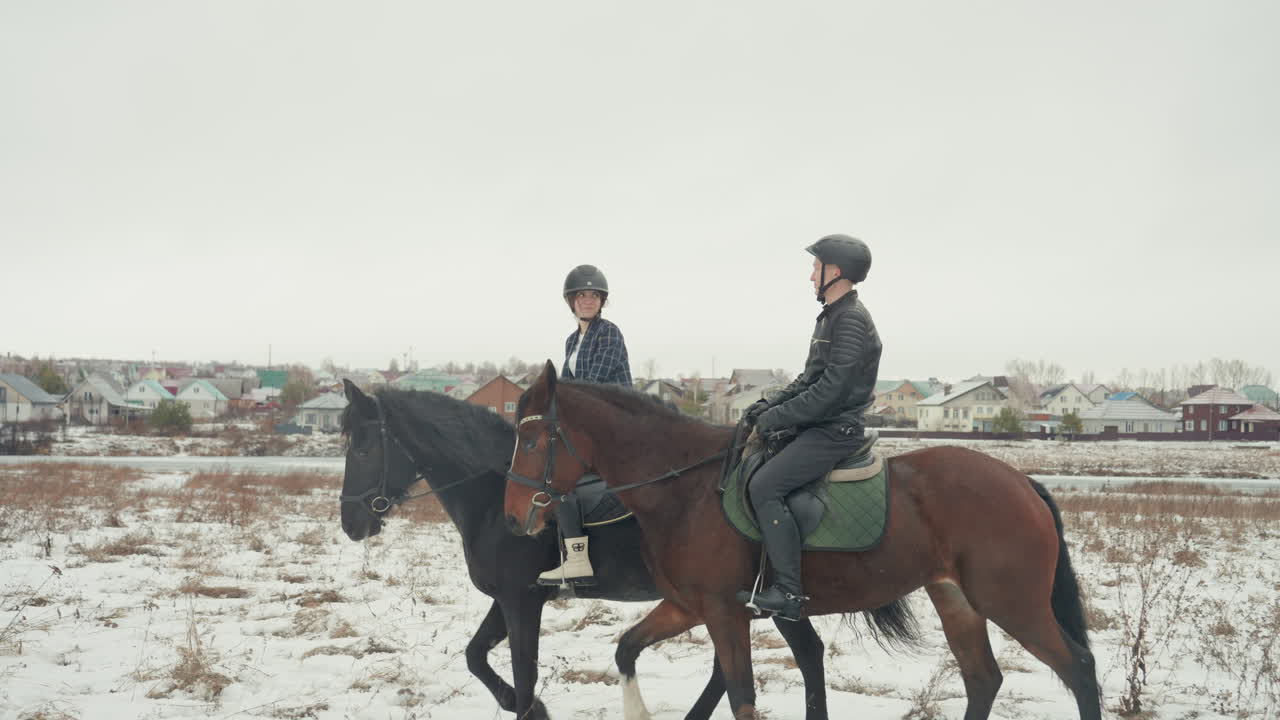 Due cavalieri caucasici su un campo innevato, istruttore che guida lo studente attraverso una lezione di equitazione in tandem, trotto lento attraverso la campagna invernale, skyline del villaggio in lontananza, toni tenui, momento di coaching sereno.