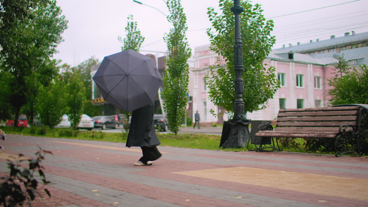 Pretty woman in long black coat walking on city sidewalk spinning umbrella near bench on cold windy day with cars, trees, urban buildings and moody street atmosphere thoughtful seasonal scene