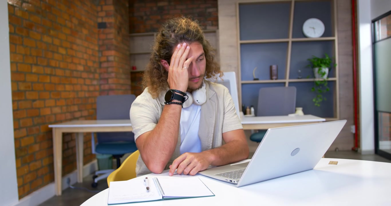 Uomo seduto a un tavolo rotondo bianco in un ufficio di coworking, lavorando su un laptop argentato e massaggiandosi la tempia.