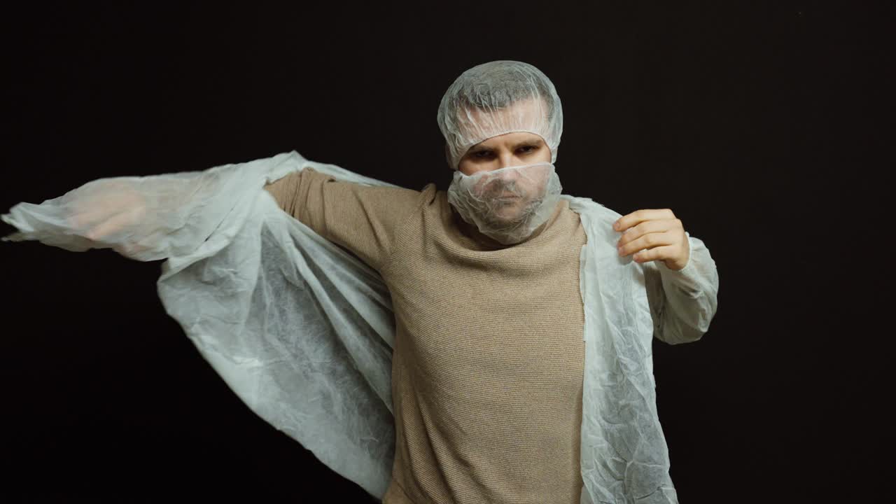 Muž si obléká jednorázový ochranný plášť, pokrývku hlavy a obličejovou masku na tmavém pozadí. Koncept osobních ochranných prostředků, hygienických norem a bezpečnostních postupů v lékařství a pracovním prostředí.