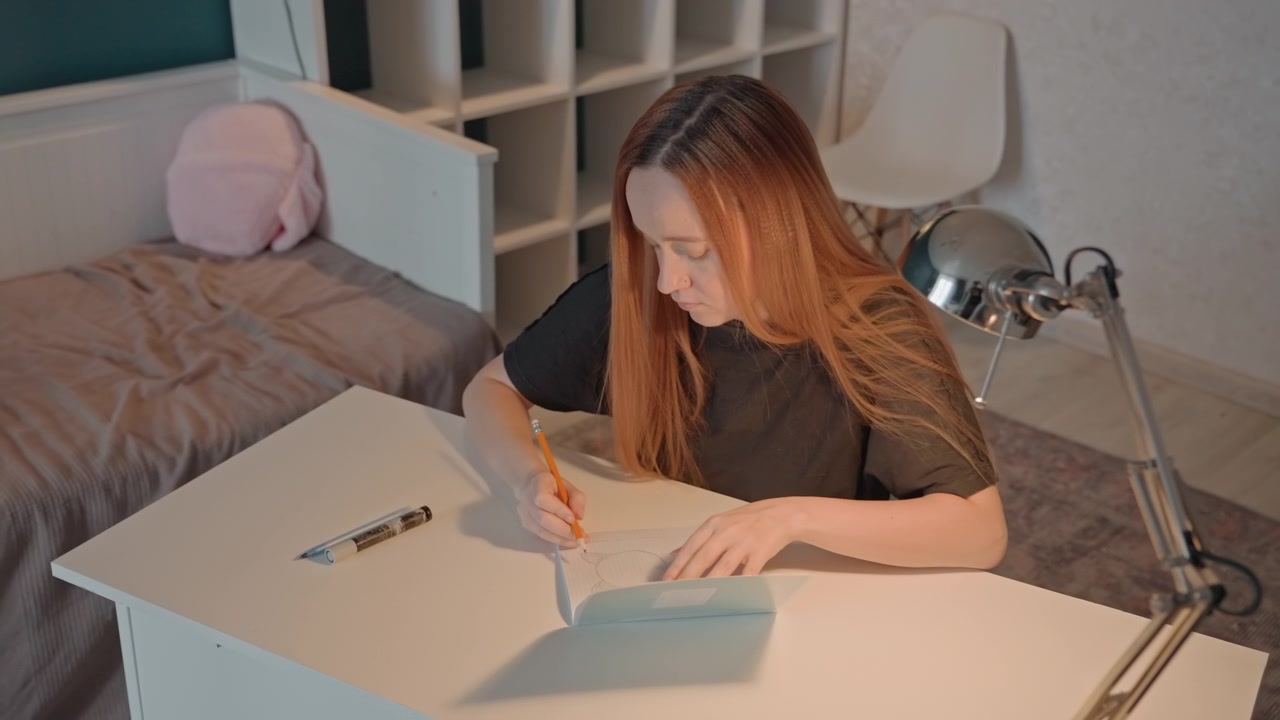 Young woman with long reddish brown hair draws at white desk. Woman wears black top and uses graph paper on table alongside. Behind her is bed with gray bedding and pink plush toy