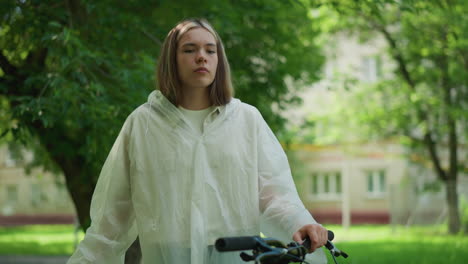 young woman wearing a translucent raincoat stands lost in thought, hand resting on bicycle handlebar, surrounded by greenery and trees, she gazes contemplatively into the distance