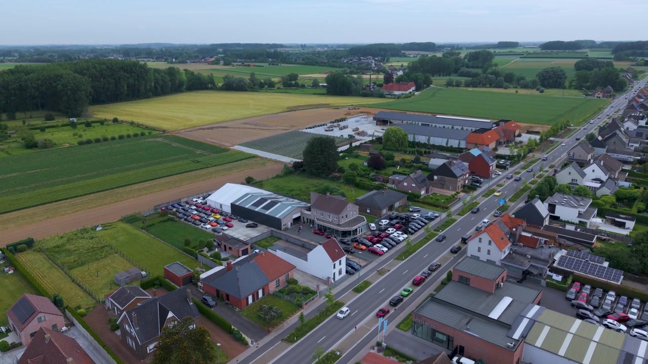 De drone trekt langzaam terug van een autodealer om de omliggende stad, landbouwgrond en het wegennet te onthullen. Een breed luchtbeeld legt het lokale landschap bij daglicht vast.