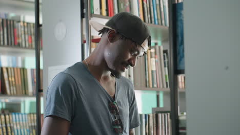 young man wearing cap and sunglasses smiling while reading book near shelf in bright library, surrounded by rows of colorful books, creating warm and relaxed atmosphere of learning and curiosity