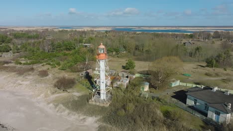 Aerial-establishing-view-of-white-colored-Pape-lighthouse,-Baltic-sea-coastline,-Latvia,-white-sand-beach,-large-waves-crashing,-sunny-day-with-clouds,-wide-revealing-drone-shot-moving-backward