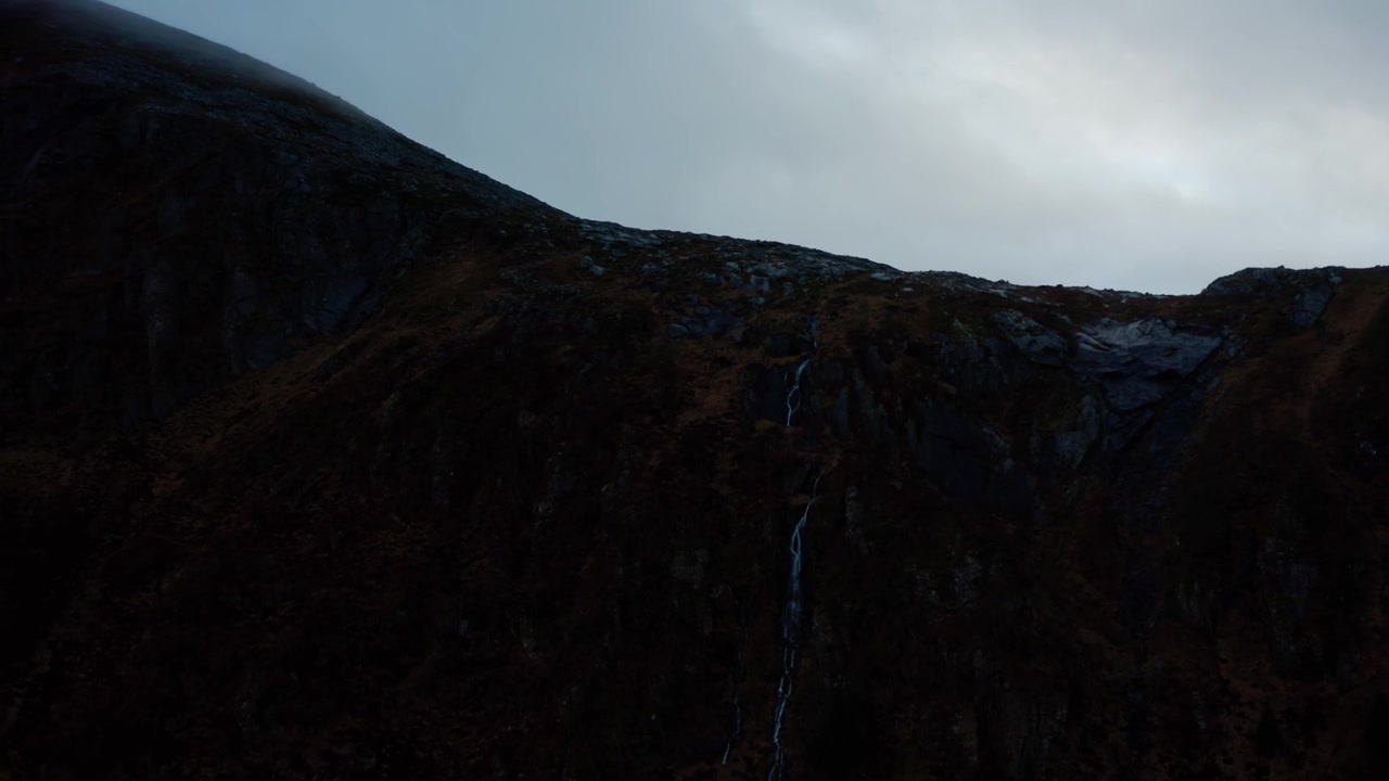 Close-up of a small waterfall on a mountain flowing through rocks and vegetation, panning out to reveal a mist-covered summit and a wide mountain range under a dark gray, cloudy sky.