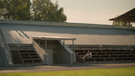 woman seated alone on stadium bleachers under metal roof, appearing deep in thought. the serene background includes lush green trees and gentle sunlight