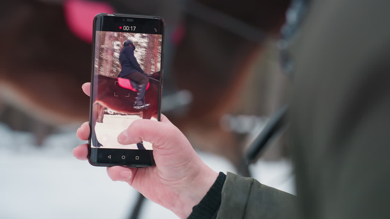 Un hombre graba una escena ecuestre al aire libre, un fotógrafo capta a un jinete y a su caballo con la lente de un smartphone, una persona toma una fotografía de cerca de un jinete a caballo en un claro nevado del bosque con un dispositivo móvil